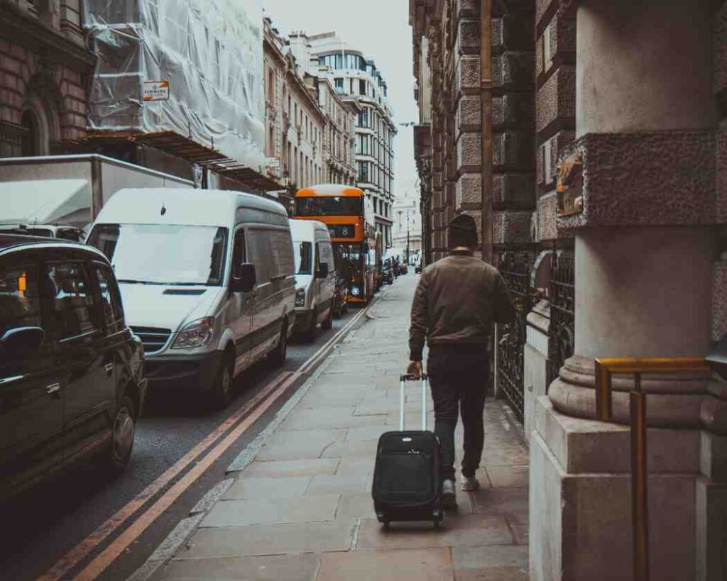 A long-term traveller walking with a suitcase on a busy city sidewalk, featuring classic European architecture, a double-decker bus, and urban traffic.